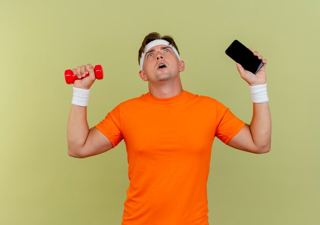 Impressionato giovane bell'uomo sportivo che indossa la fascia e braccialetti tenendo il telefono cellulare e il manubrio guardando in alto isolato su verde oliva