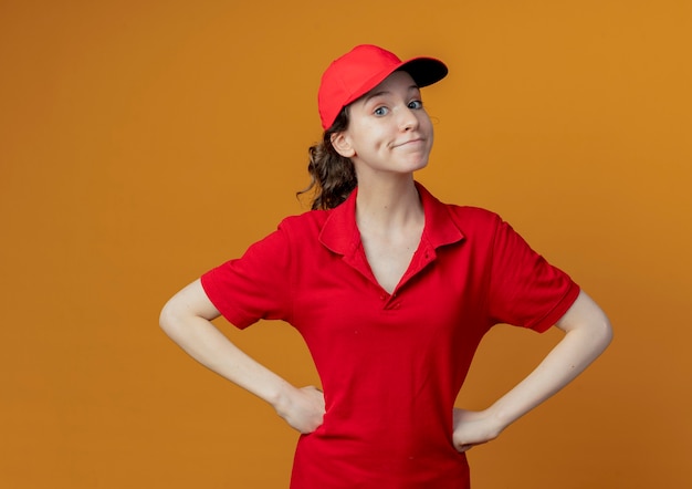 Impressionato giovane bella consegna ragazza in uniforme rossa e berretto mettendo le mani sulla vita guardando la telecamera isolata su sfondo arancione con spazio di copia