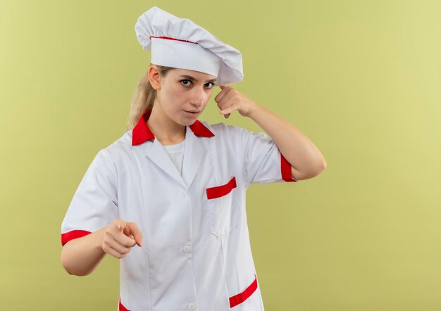 Impressionato giovane bella cuoca in uniforme da chef che fa un gesto di chiamata e indica isolata sulla parete verde con spazio di copia