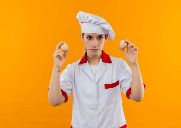 Impressionato giovane bella cuoca in uniforme da chef che tiene le uova isolate sulla parete arancione