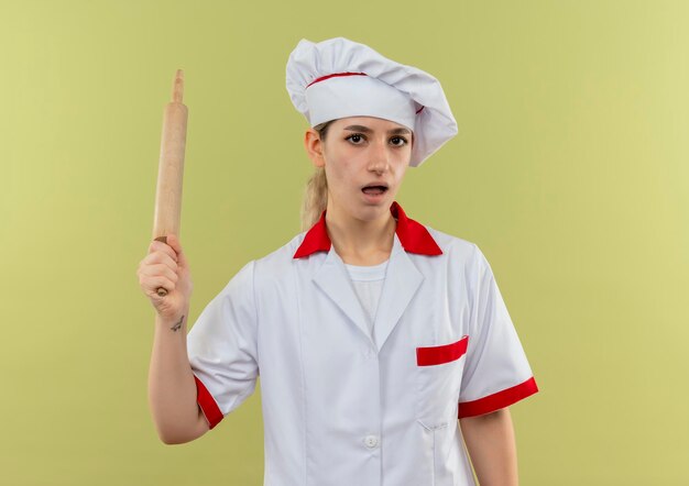 Impressionato giovane bella cuoca in uniforme da chef che tiene mattarello isolato sulla parete verde