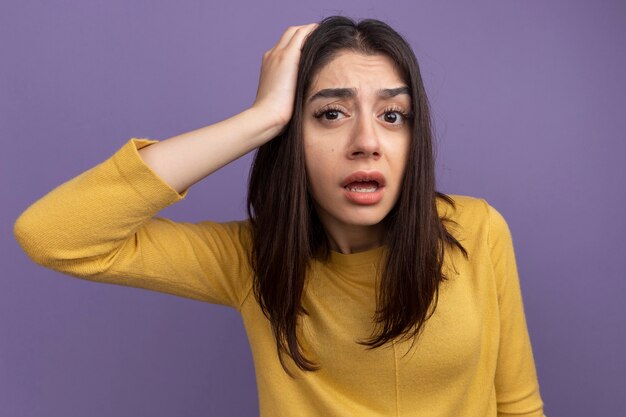 Impressionato giovane bella donna che guarda davanti mettendo la mano sulla testa isolata sul muro viola