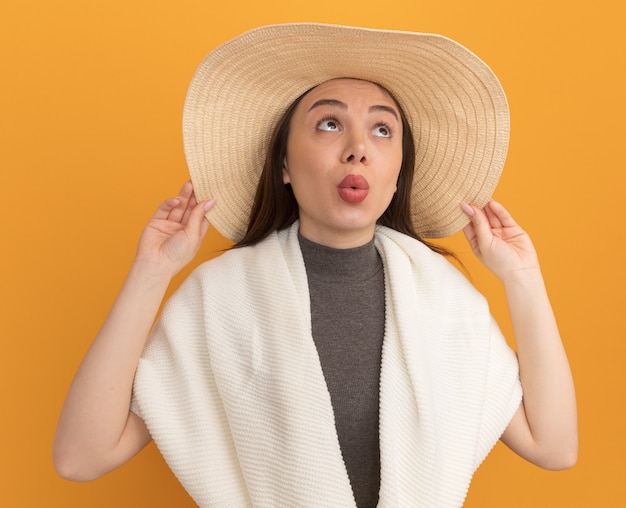 Impressionato giovane bella donna che indossa il cappello da spiaggia che afferra il cappello guardando in alto con le labbra increspate