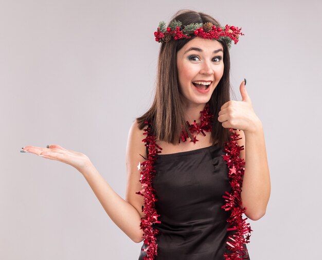 Impressionato giovane bella ragazza caucasica che indossa ghirlanda di testa di Natale e ghirlanda di orpelli intorno al collo che mostra la mano vuota e il pollice alzato guardando la telecamera isolata su sfondo bianco