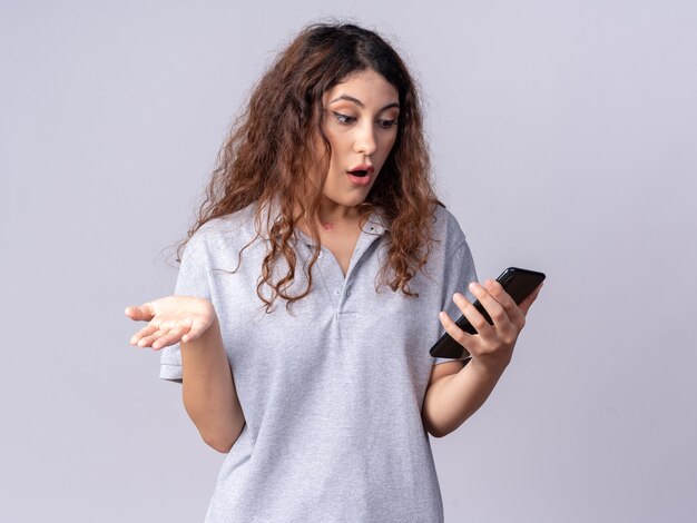 Impressionato giovane bella ragazza caucasica che tiene e guarda il telefono cellulare che mostra la mano vuota isolata sul muro bianco con spazio per le copie