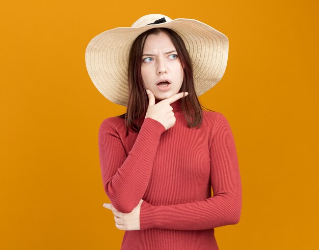 Impressionato giovane bella ragazza che indossa cappello da spiaggia tenendo la mano sul mento guardando a lato