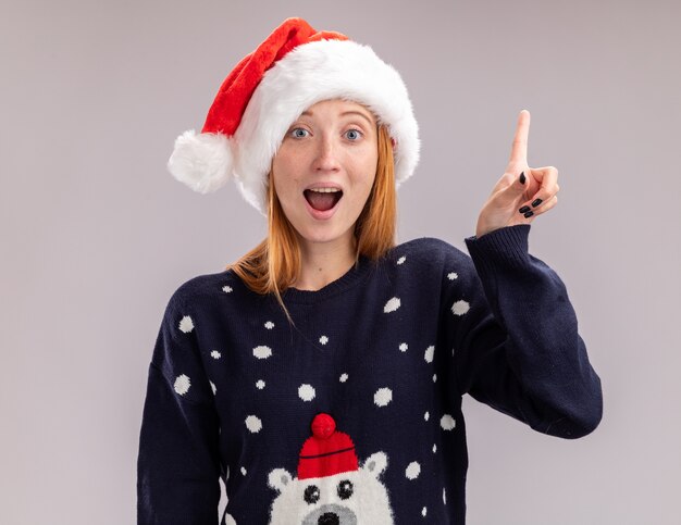 Impressionato giovane bella ragazza che indossa il cappello di Natale indica in alto isolato sul muro bianco