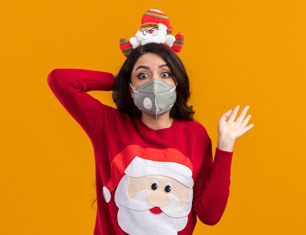 impressionato giovane bella ragazza che indossa la fascia di Babbo Natale e maglione con maschera protettiva tenendo la mano dietro la testa guardando la telecamera che mostra la mano vuota isolata su sfondo arancione