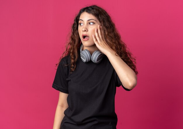 Impressionato giovane bella ragazza che indossa le cuffie sul collo tenendo la mano sul viso guardando il lato isolato sul muro cremisi con spazio copia