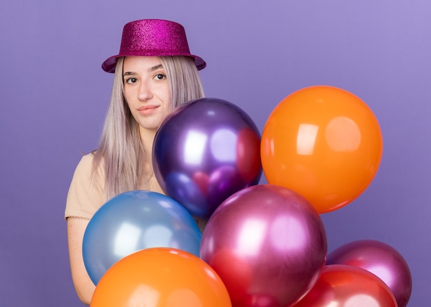 Impressionato giovane bella ragazza che indossa un cappello da festa in piedi dietro palloncini isolati sulla parete blu