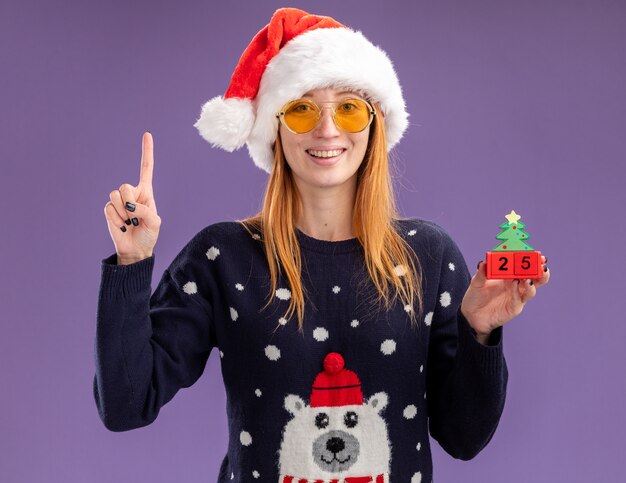Impressionato giovane bella ragazza che indossa un maglione di Natale e cappello con gli occhiali che tengono i punti del giocattolo di Natale in alto isolato su sfondo viola
