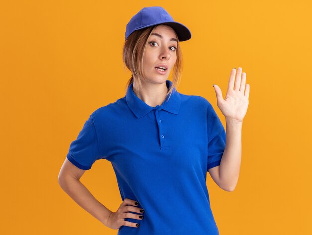 Impressionato giovane bella ragazza di consegna in stand uniforme con la mano alzata sull'arancio