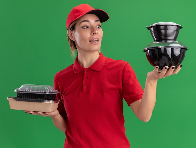 Impressionato giovane bella ragazza di consegna in uniforme tiene i pacchetti di cibo e guarda i contenitori di cibo sul verde