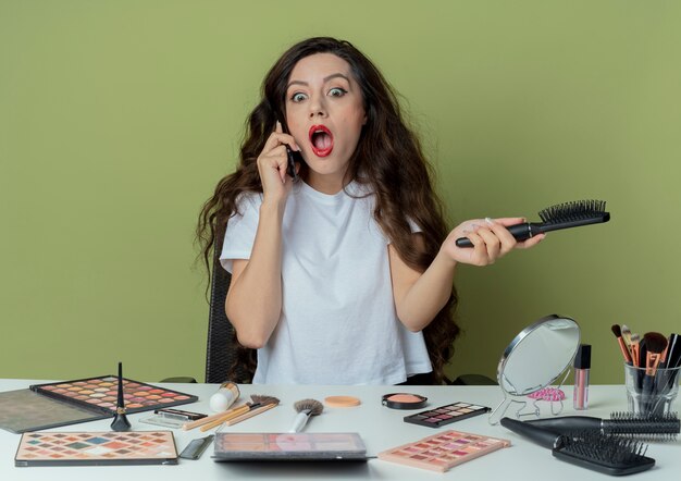 Impressionato giovane bella ragazza seduta al tavolo per il trucco con strumenti di trucco tenendo il pettine e parlando al telefono guardando in basso isolato su sfondo verde oliva