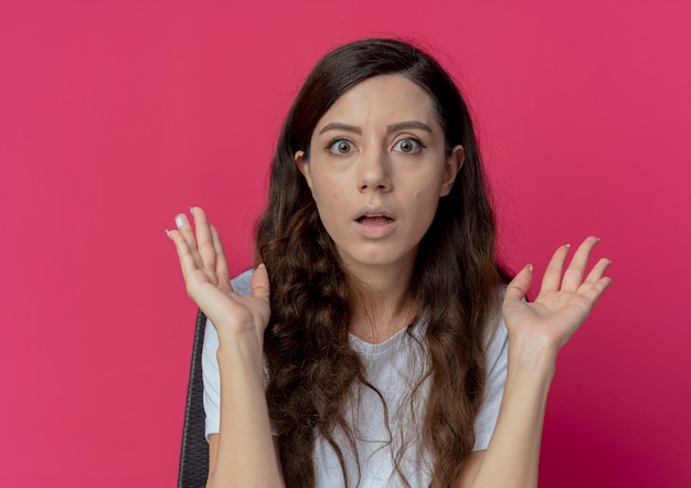 Impressionato giovane bella ragazza seduta al tavolo per il trucco con strumenti per il trucco che mostra le mani vuote guardando la telecamera isolata su sfondo cremisi