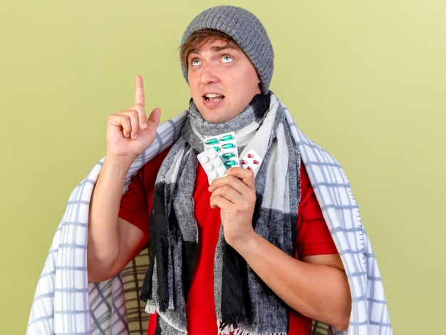 Impressionato giovane bello biondo malato che indossa cappello invernale e sciarpa avvolto in plaid che tiene confezioni di pillole mediche guardando e rivolto verso l'alto isolato sulla parete verde oliva