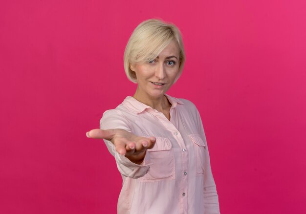 Impressionato giovane bionda donna slava allungando la mano verso la telecamera isolata su sfondo rosa con copia spazio