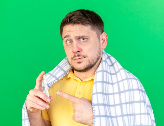 Impressionato giovane biondo malato uomo slavo avvolto in plaid tiene e punta al termometro isolato sulla parete verde con copia spazio