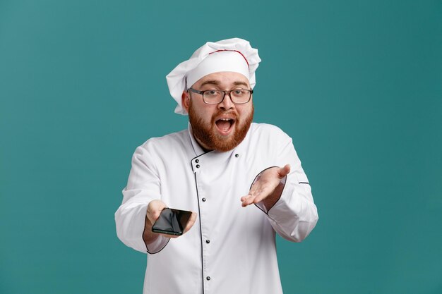 Impressionato giovane chef maschio che indossa occhiali uniformi e cappuccio guardando la fotocamera allungando il telefono cellulare verso la fotocamera che mostra la mano vuota isolata su sfondo blu