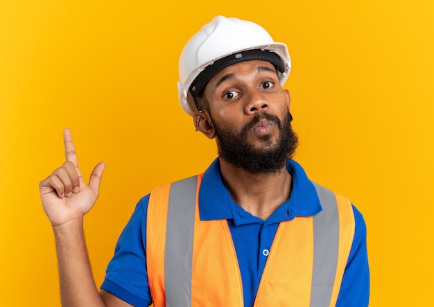 impressionato giovane costruttore uomo in uniforme con casco di sicurezza rivolto verso l'alto isolato sulla parete arancione con copia spazio