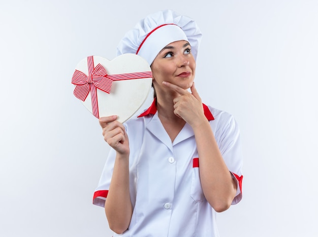 Impressionato giovane cuoca che indossa l'uniforme dello chef tenendo la scatola a forma di cuore afferrato il mento isolato sul muro bianco