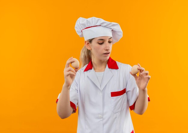 Impressionato giovane cuoco carino in uniforme da chef che tiene e guarda le uova isolate sulla parete arancione orange