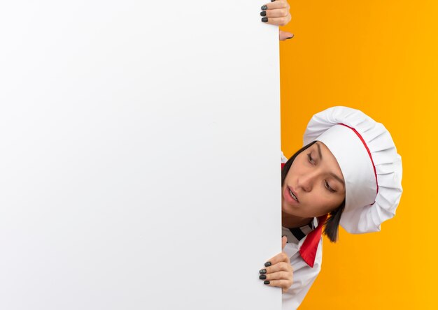 Impressionato giovane cuoco femminile in uniforme da chef giovane cuoco femminile in uniforme da chef tenendo e guardando da dietro il muro bianco alla parete isolata su con lo spazio della copia