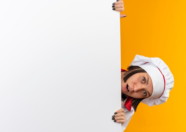 Impressionato giovane cuoco femminile in uniforme da chef giovane cuoco femminile in uniforme da chef tenendo e guardando da dietro il muro bianco isolato su con lo spazio della copia