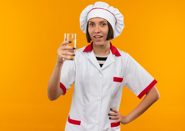 Impressionato giovane cuoco femminile in uniforme del cuoco unico che tiene il bicchiere d'acqua mettendo la mano sulla vita isolata sull'arancio con lo spazio della copia