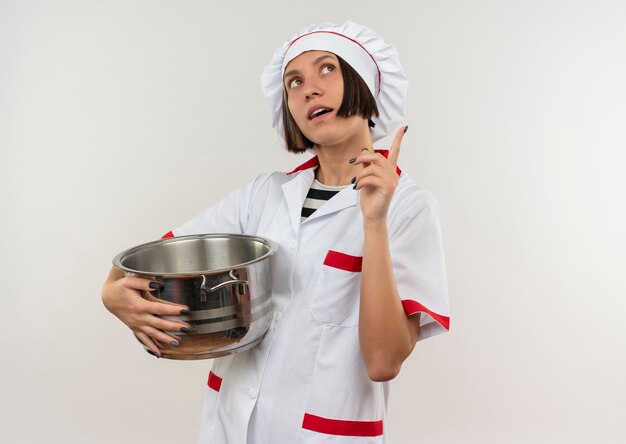 Impressionato giovane cuoco femminile in uniforme del cuoco unico che tiene la pentola che alza il dito e che osserva in su isolato su bianco con lo spazio della copia