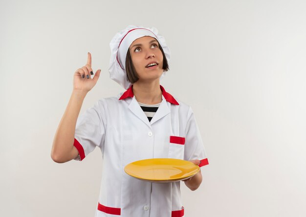 Impressionato giovane cuoco femminile in uniforme del cuoco unico che tiene piatto che osserva e che indica su isolato su bianco con lo spazio della copia