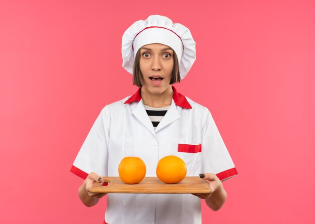 Impressionato giovane cuoco femminile in uniforme del cuoco unico che tiene tagliere con arance su di esso isolato su rosa con spazio di copia