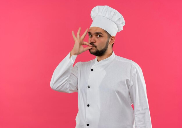 Impressionato giovane cuoco maschio in uniforme da chef che fa un gesto gustoso isolato sulla parete rosa con spazio per le copie