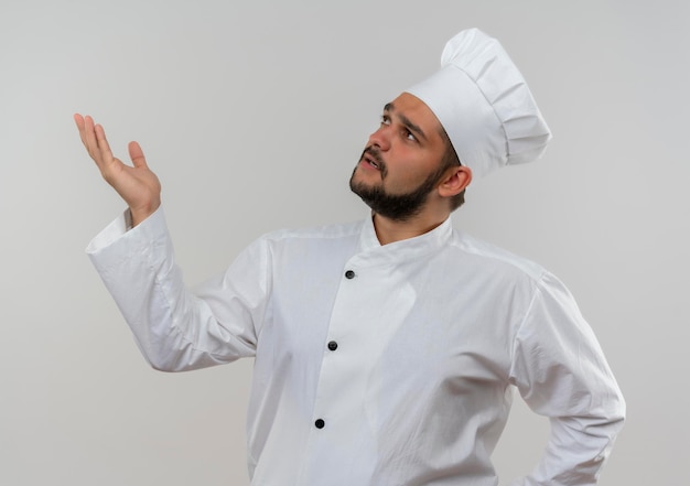 Impressionato giovane cuoco maschio in uniforme da chef che guarda in alto e mostra la mano vuota isolata sul muro bianco