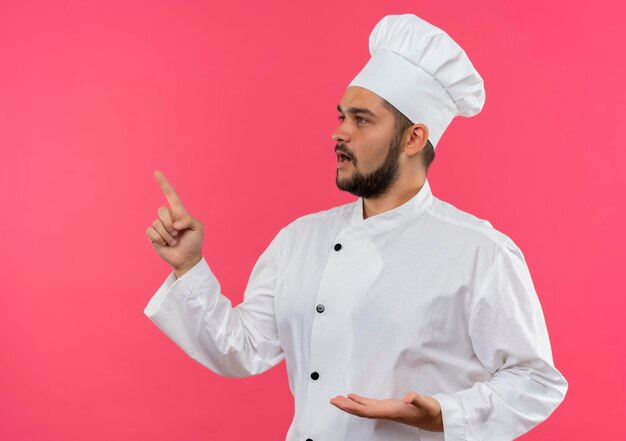 Impressionato giovane cuoco maschio in uniforme da chef che mostra la mano vuota che guarda e indica il lato isolato sulla parete rosa con spazio di copia