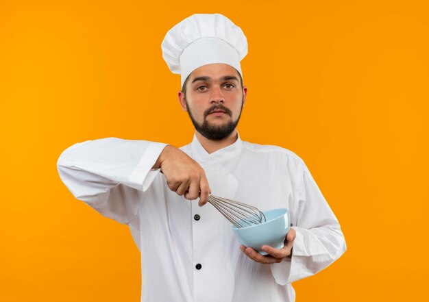 Impressionato giovane cuoco maschio in uniforme da chef che tiene in mano una frusta e una ciotola isolate sulla parete arancione orange