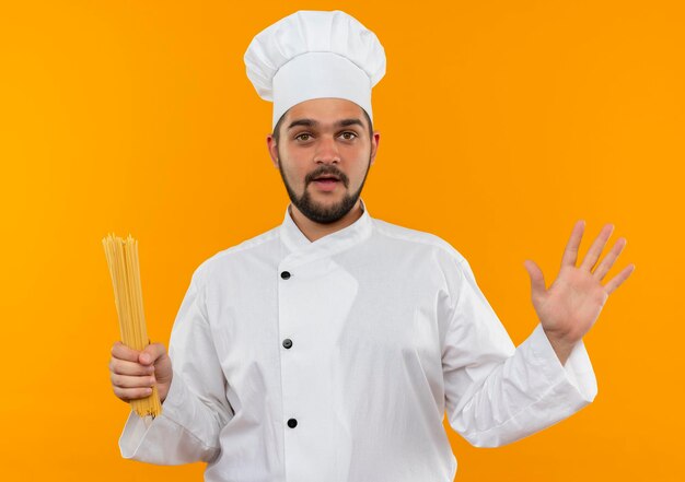 Impressionato giovane cuoco maschio in uniforme da chef che tiene la pasta degli spaghetti e mostra la mano vuota isolata sulla parete arancione