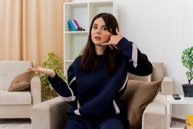 Impressionato giovane donna abbastanza caucasica seduta sulla poltrona nel soggiorno progettato facendo gesto di chiamata e mostrando la mano vuota