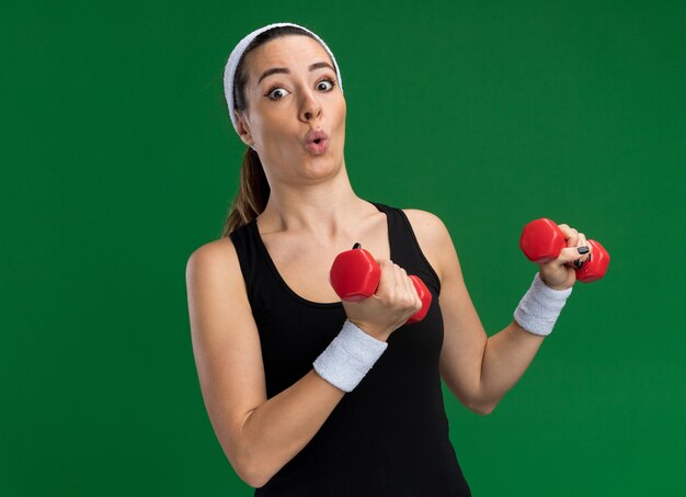 Impressionato giovane donna abbastanza sportiva che indossa fascia e braccialetti che tengono i manubri guardando la parte anteriore isolata sulla parete verde con spazio di copia