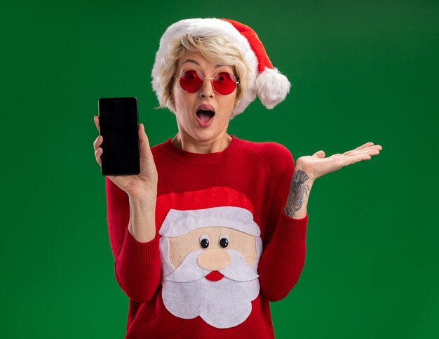 Impressionato giovane donna bionda che indossa il cappello di Natale e Babbo Natale maglione di Natale con gli occhiali che mostra il telefono cellulare alla telecamera guardando la telecamera che mostra la mano vuota isolata su sfondo verde