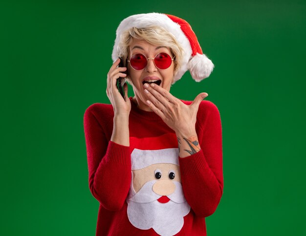 Impressionato giovane donna bionda che indossa il cappello di Natale e Babbo Natale maglione di Natale con gli occhiali parlando al telefono guardando la telecamera tenendo la mano sulla bocca isolata su sfondo verde