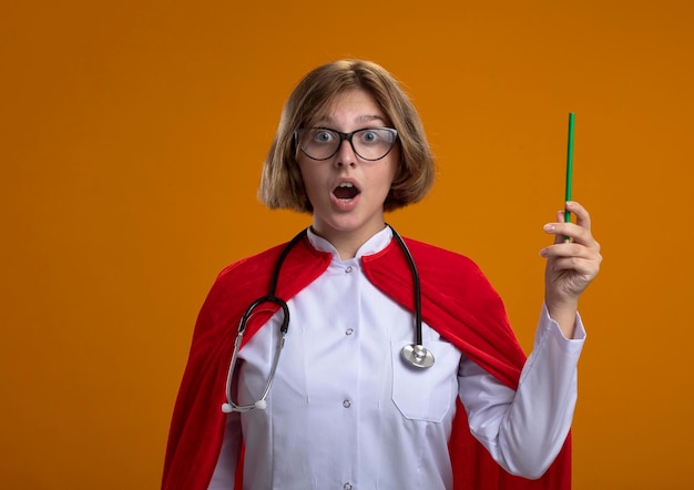 Impressionato giovane donna bionda supereroe in mantello rosso che indossa l'uniforme del medico e occhiali con lo stetoscopio che tiene la matita guardando la parte anteriore isolata sulla parete arancione con lo spazio della copia