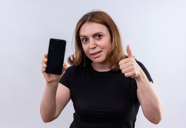 Impressionato giovane donna casuale che tiene il telefono cellulare e che mostra il pollice in su su uno spazio bianco isolato