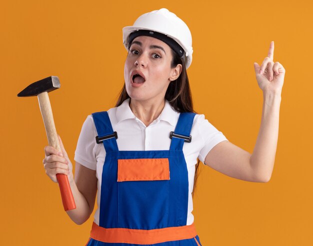 Impressionato giovane donna costruttore in uniforme che tiene i punti del martello in alto isolato sulla parete arancione