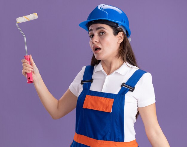 Impressionato giovane donna costruttore in uniforme che tiene mini rullo di vernice isolato sulla parete viola