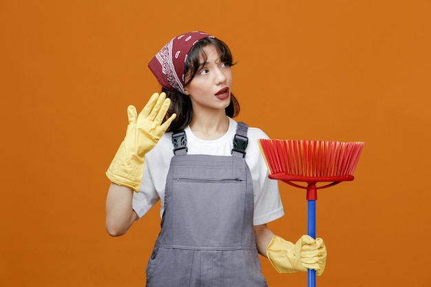 Impressionato giovane donna detergente che indossa uniforme guanti di gomma e bandana tenendo la mano in aria tenendo il mop tergipavimento guardando il lato isolato su sfondo arancione