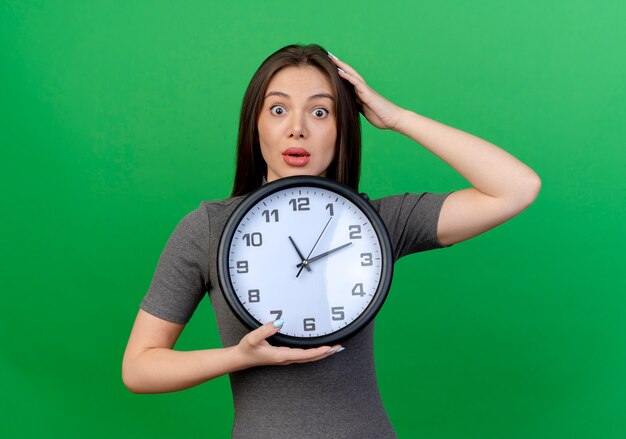 Impressionato giovane donna graziosa che tiene l'orologio e mettendo la mano sulla testa isolata su sfondo verde