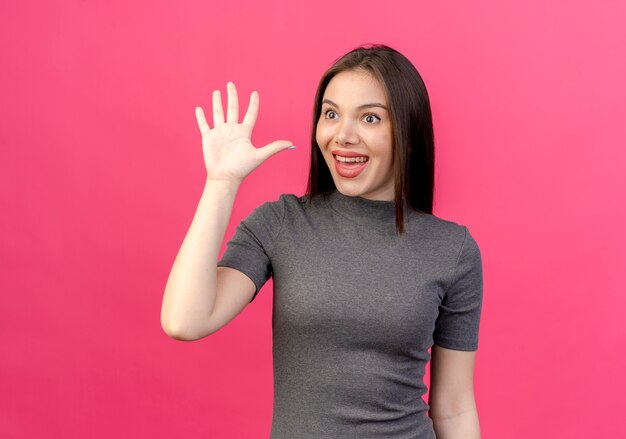 Impressionato giovane donna graziosa guardando a lato e facendo ciao gesto isolato su sfondo rosa con spazio di copia