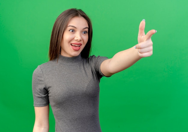 Impressionato giovane donna graziosa guardando dritto e facendo il gesto della pistola isolato su sfondo verde