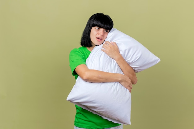 Impressionato giovane donna malata che abbraccia il cuscino guardando il lato isolato sulla parete verde oliva